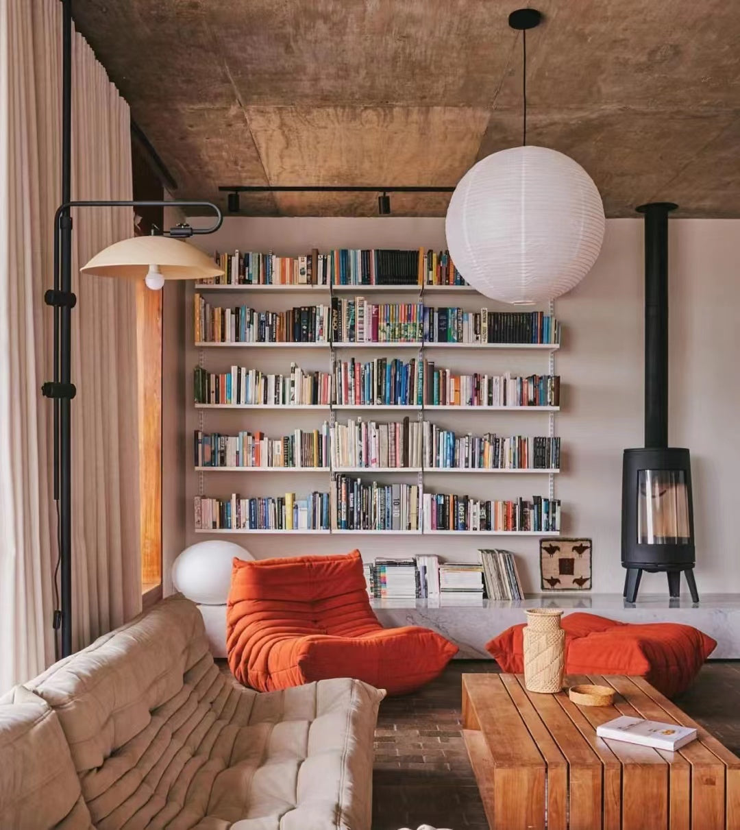 A cozy living room with a large bookshelf, orange lounge chairs, a wooden coffee table, and a modern fireplace, creating a warm and stylish reading nook.