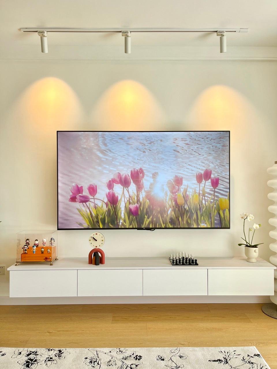 A modern living room with a wall-mounted TV showing tulips by the water, white console with decor, and soft lighting, creating a fresh clean vibe in spring.
